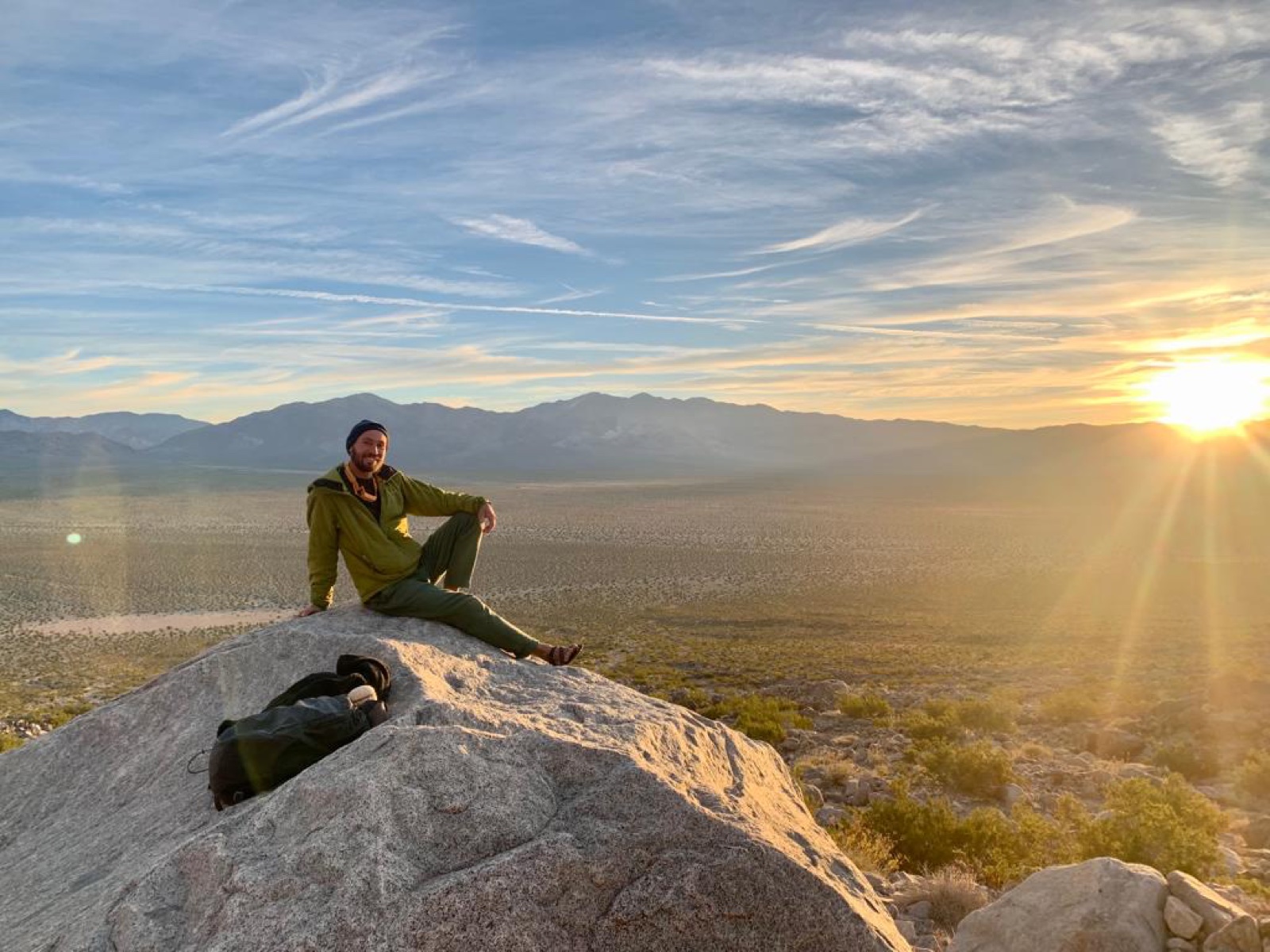 The author seated on a sunlit desert boulder with mountains beyond